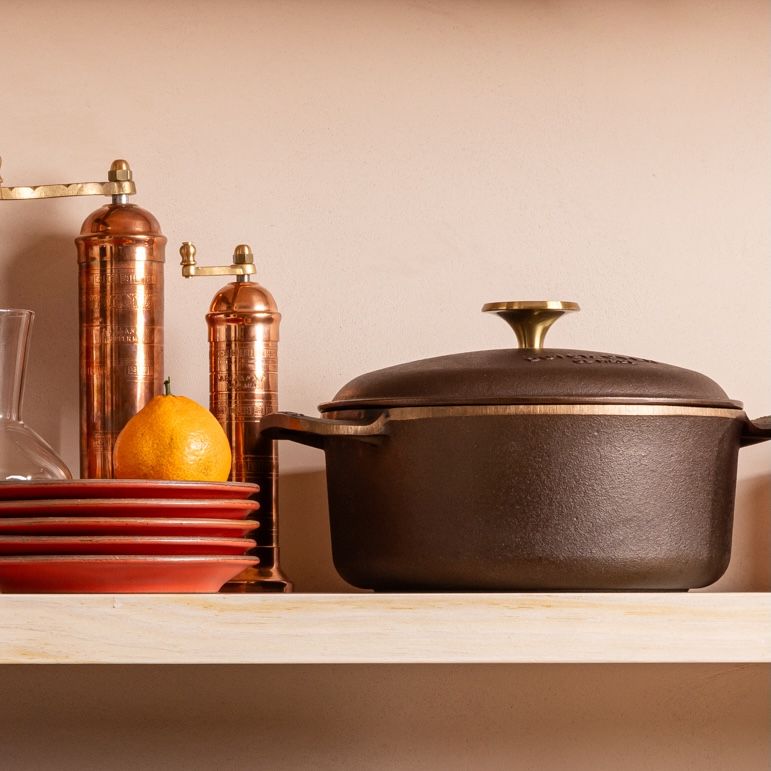 A floating shelf has a large cast iron dutch oven sitting on it. Also on the shelf are copper greek pepper and salt mills, a stack of red ceramic plates with an orange on top.
