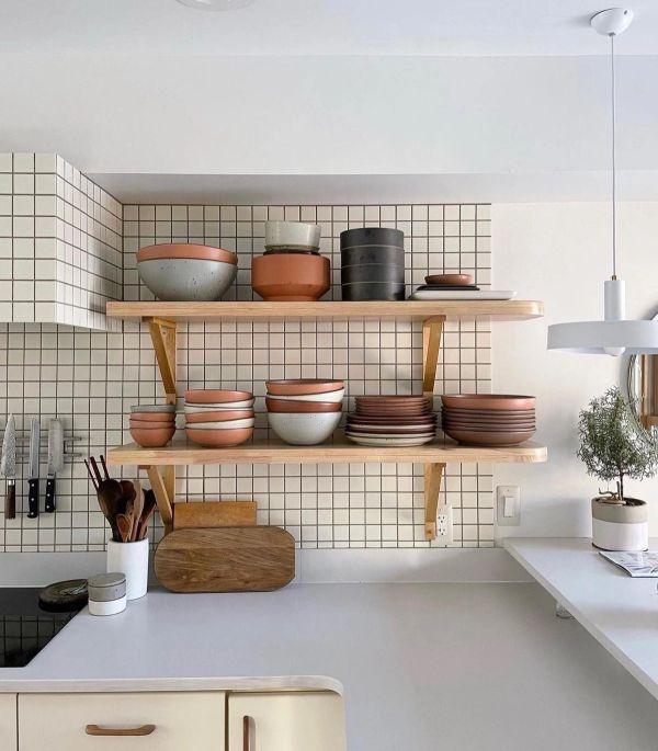 A mostly white, modern kitchen with a tight subway tile backsplash that extends to the ceiling. Exposed wooden shelving displays a collection of East Fork pottery.