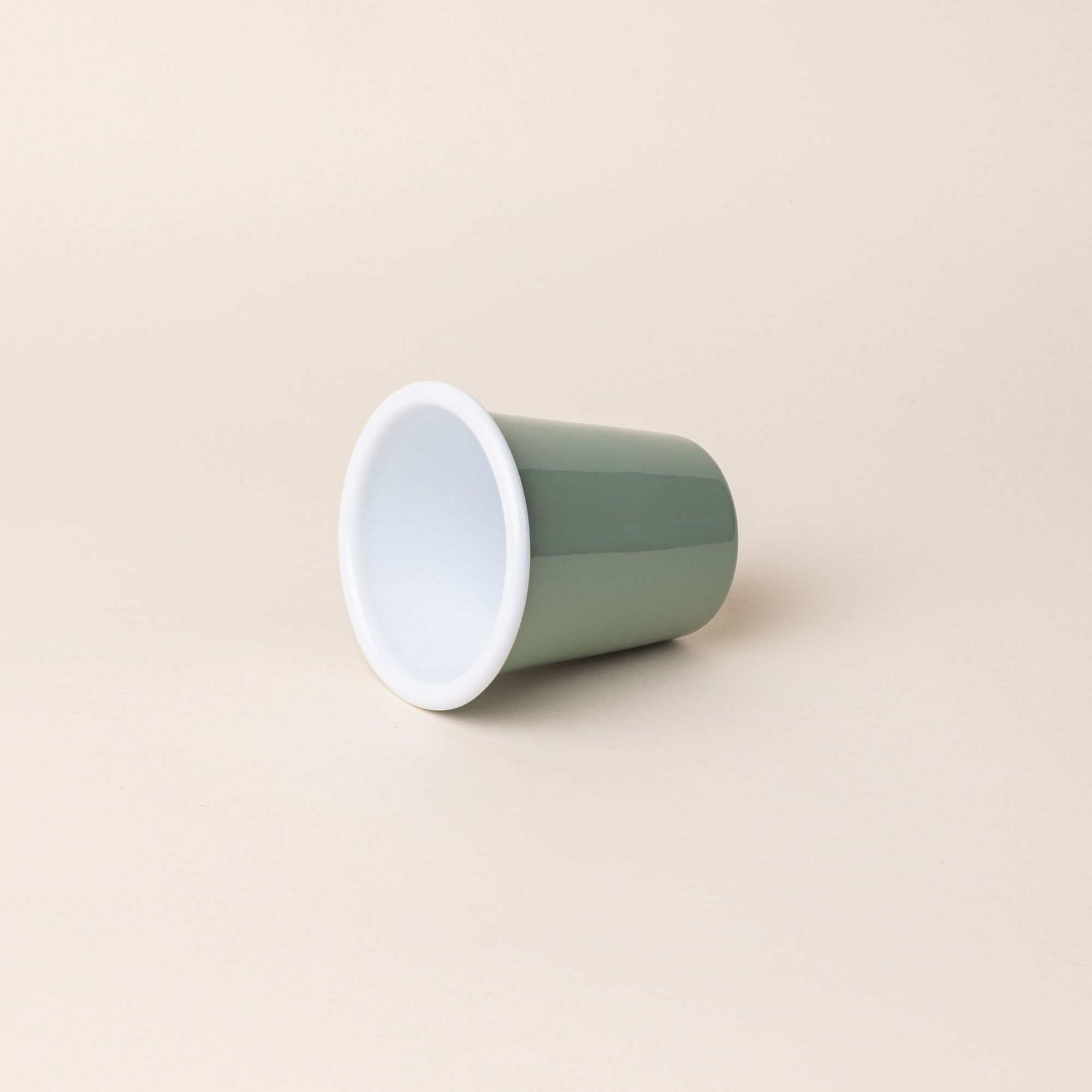 A short enamel cup with a sage green exterior and white interior sitting on its side.