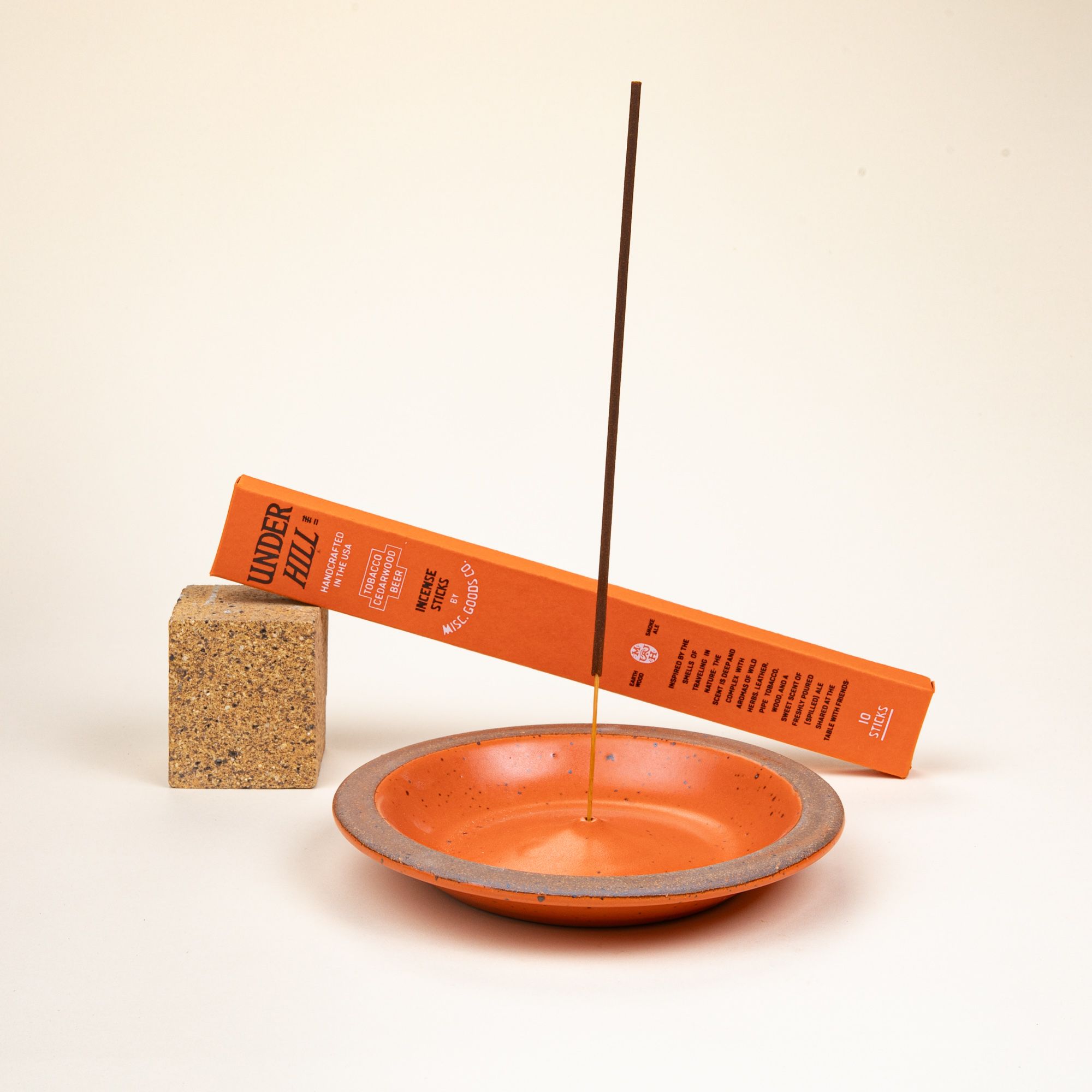 An incense stick sits in a ceramic plate-shaped incense stick holder with an unglazed rim and glazed in a bold orange color featuring lots of iron speckles. Behind the holder is a propped-up rectangular box of incense sticks.