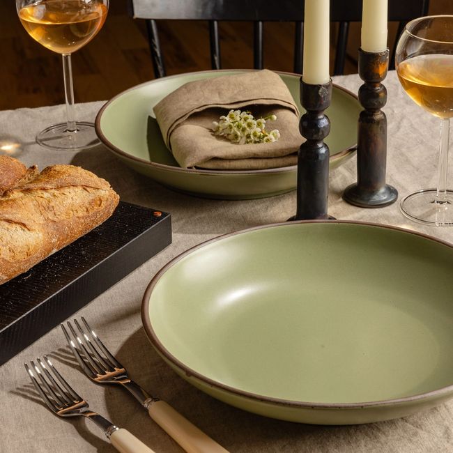 A dining table set with ceramic couple plates in a calm sage green, taper candles, and a glass of wine.