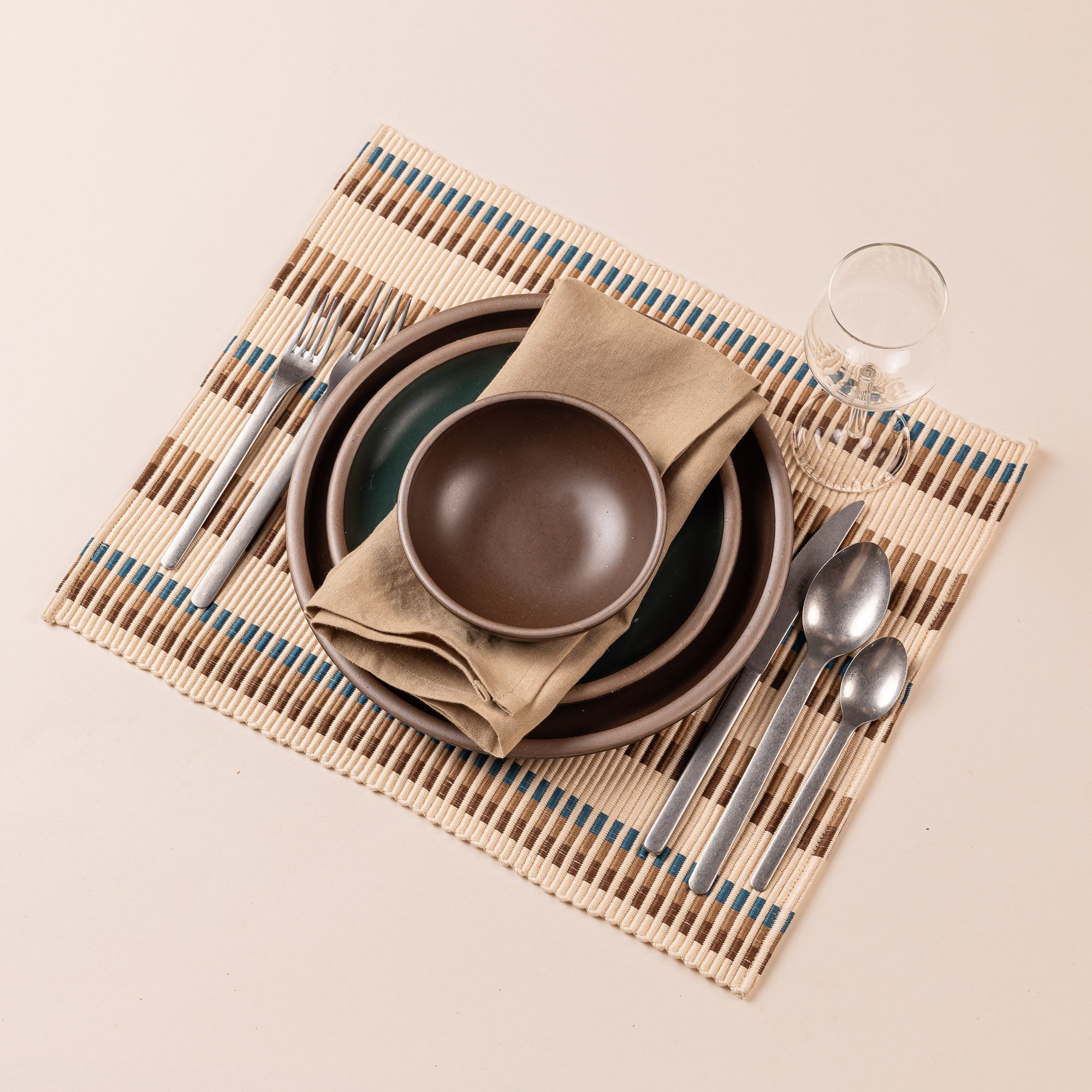 A table setting featuring a stack of brown and dark teal plates and bowls, light brown napkin, silver minimal flatware, and a wine glass all sitting on a rectangle placemat. The placemat has an offset stripe design that is hand woven in cream, brown, tan, and blue colors.