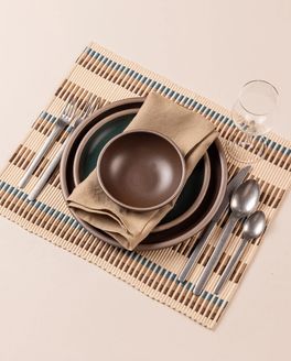 A table setting featuring a stack of brown and dark teal plates and bowls, light brown napkin, silver minimal flatware, and a wine glass all sitting on a rectangle placemat. The placemat has an offset stripe design that is hand woven in cream, brown, tan, and blue colors.