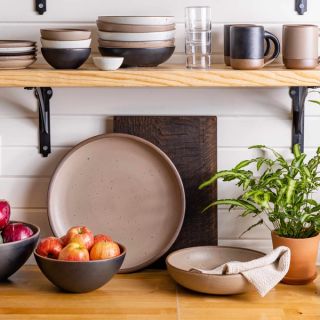 In a kitchen setting, there are wooden shelves holding ceramic bowls and mugs in a graphite, muted tan, and white colors, along with a set of stacking water glasses. Below the shelf is a counter filled with larger ceramic pieces in a serving platter and large fruit bowl along with a plant, and a large shallow bowl holding linen napkins.