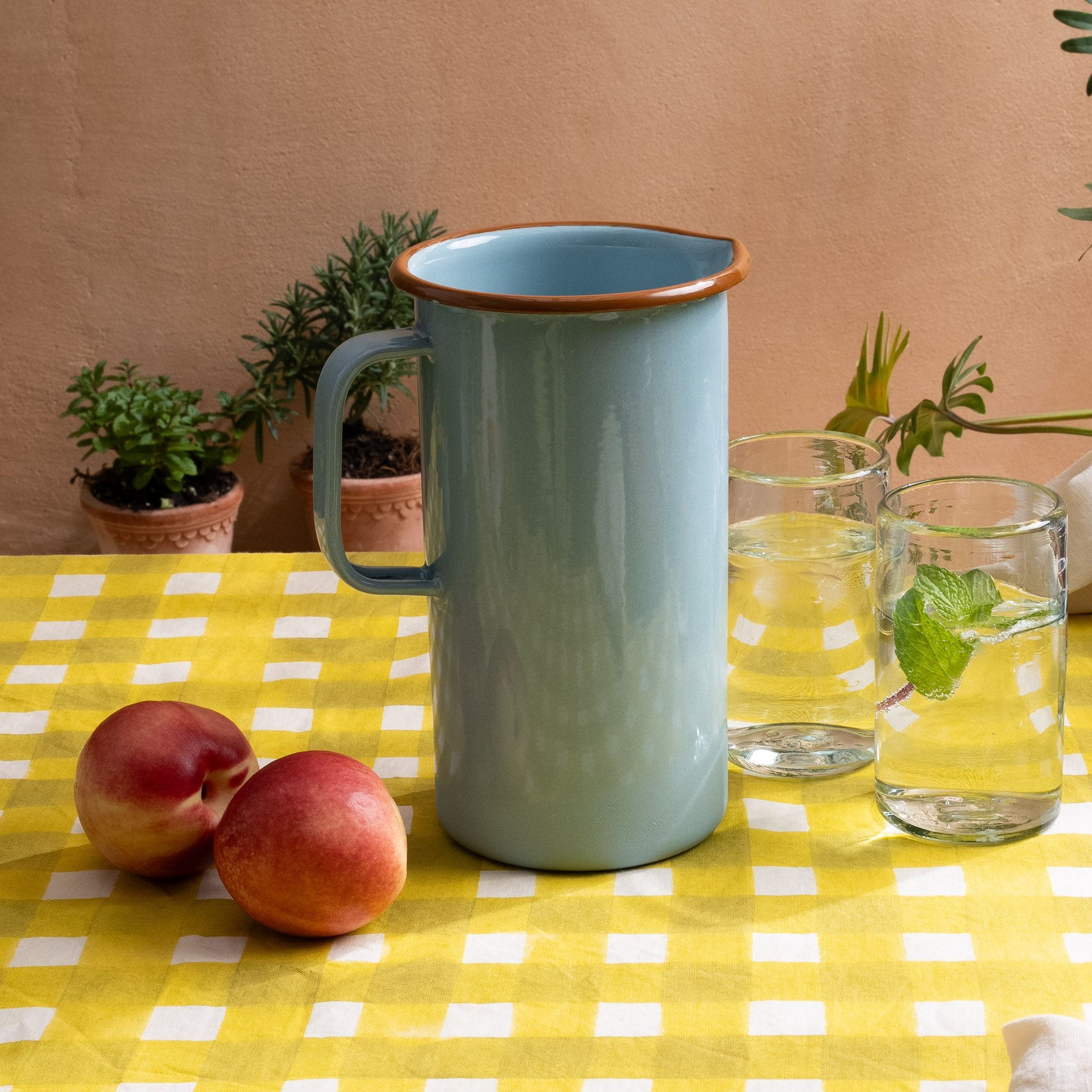 A large enamel pitcher in a robin's egg blue sitting on a bright yellow crosshatch tablecloth with tall clear glasses, and apples.