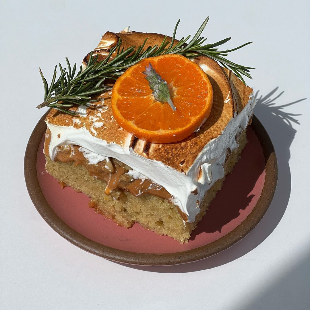 A square slice of cake topped with meringue, orange slice, and rosemary on a pink ceramic plate