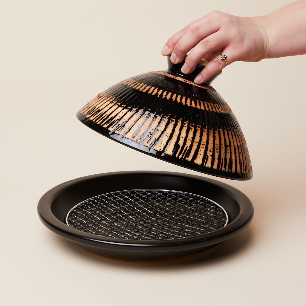 A dark clay cooking vessel comprised of a flat plate with metal grill and a cone-shaped lid
