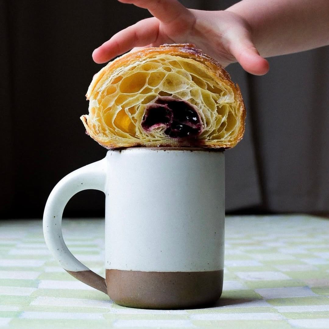 Photo by @nolalogan_. A hand grabbing a chocolate croissant, sitting on top of a medium sized ceramic mug with handle in a cool white color featuring iron speckles and unglazed rim and bottom base.