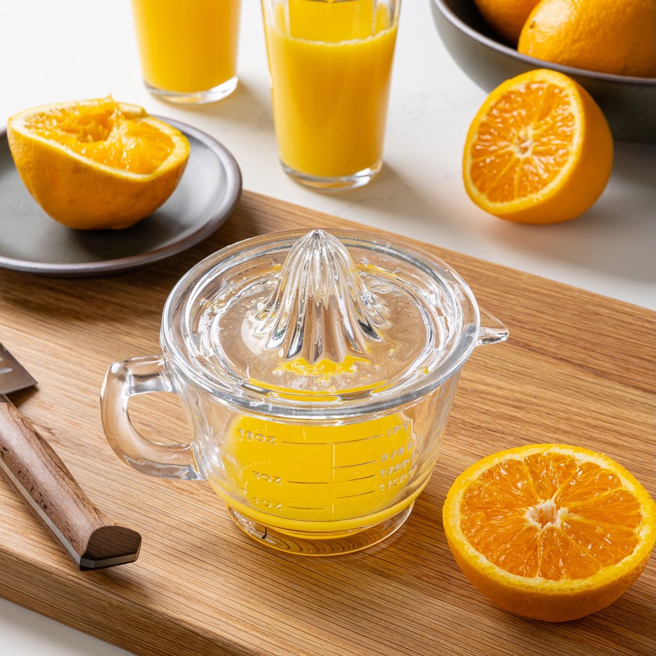 A traditional glass juicer with a handle and juice inside, surrounded by slices of oranges, a knife, and glasses filled with orange juice.