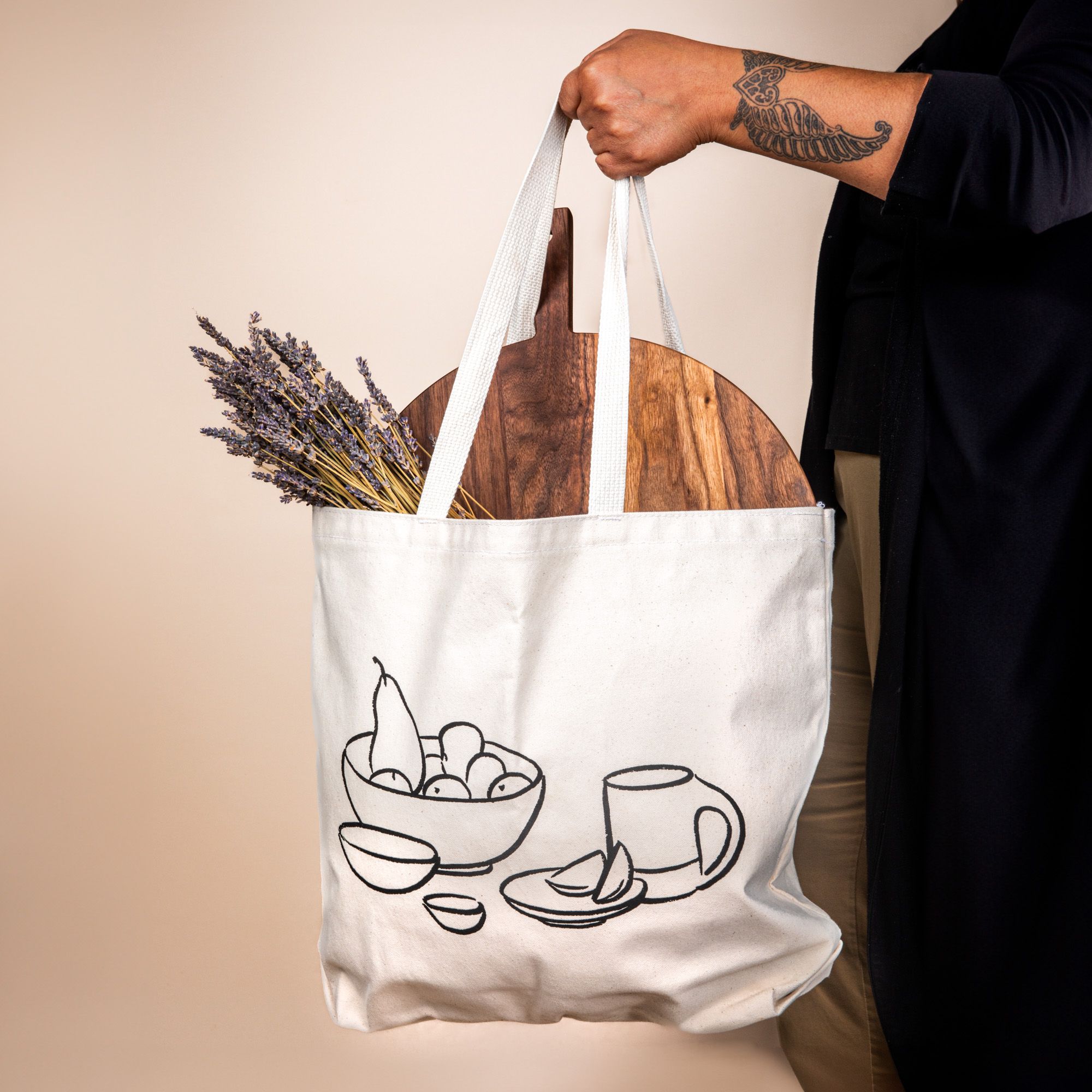 Person holding a cream canvas tote on their shoulder with their hand holding onto the handle - the tote has a charcoal illustration on the front of a bowl with fruit, little bowls, plate, and a mug. Underneath, it reads 'East Fork is a vessel'.