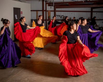 In a rehearsal space, there are young girls dancing with large colorful long skirts in red, purple, and orange colors.