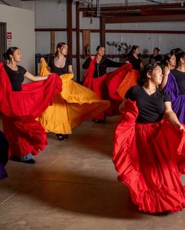 In a rehearsal space, there are young girls dancing with large colorful long skirts in red, purple, and orange colors.
