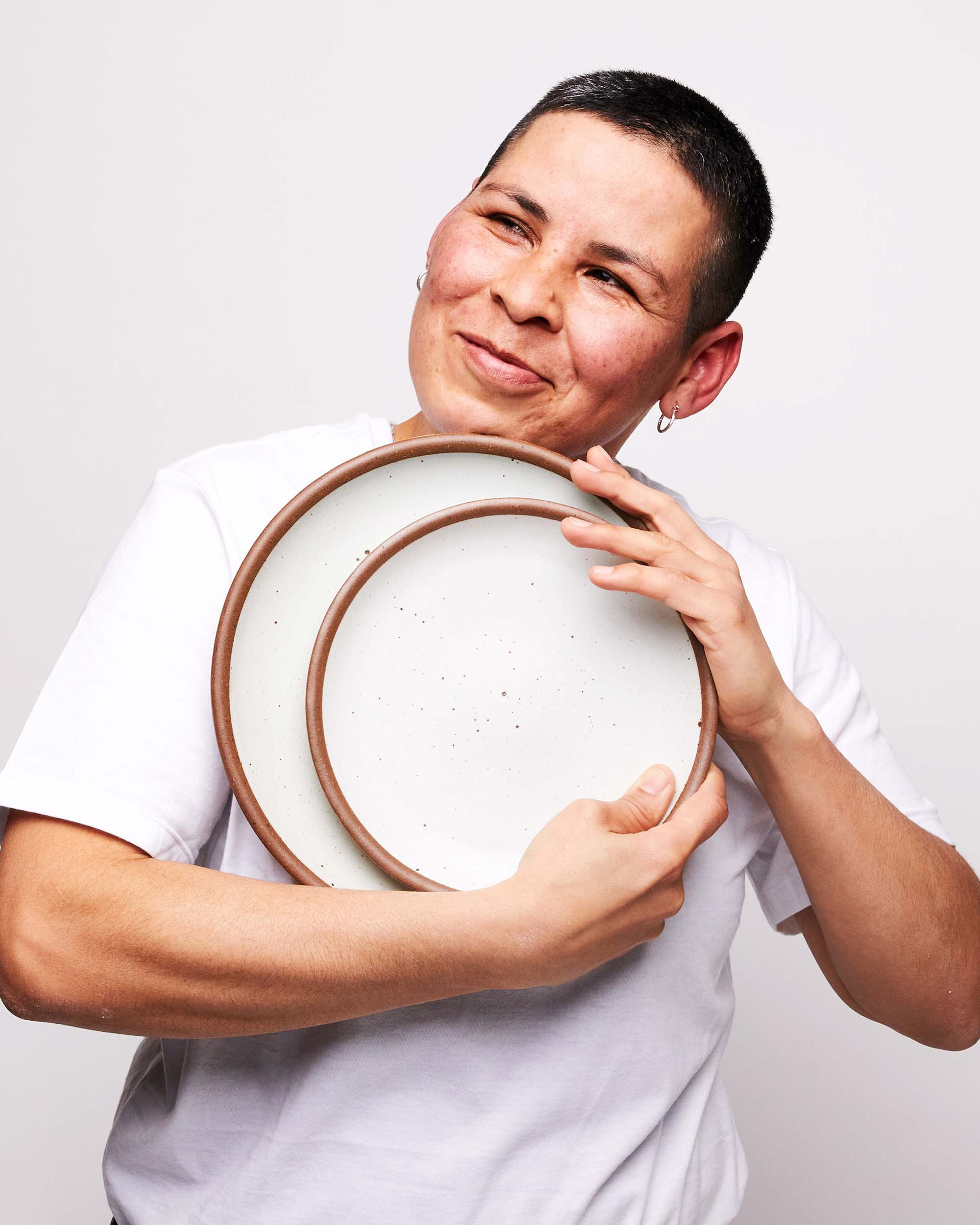 A person holds ceramic plates in a cool white color close to the chest and smiles off to the side.