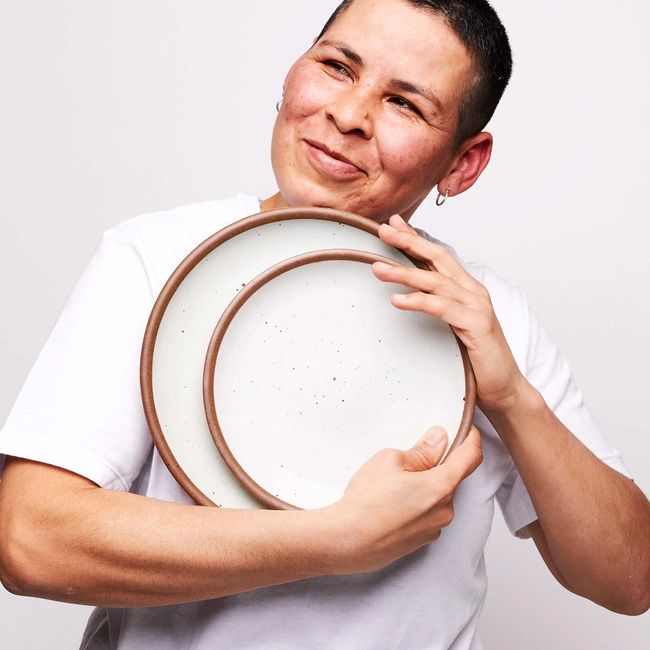 A person holds ceramic plates in a cool white color close to the chest and smiles off to the side.