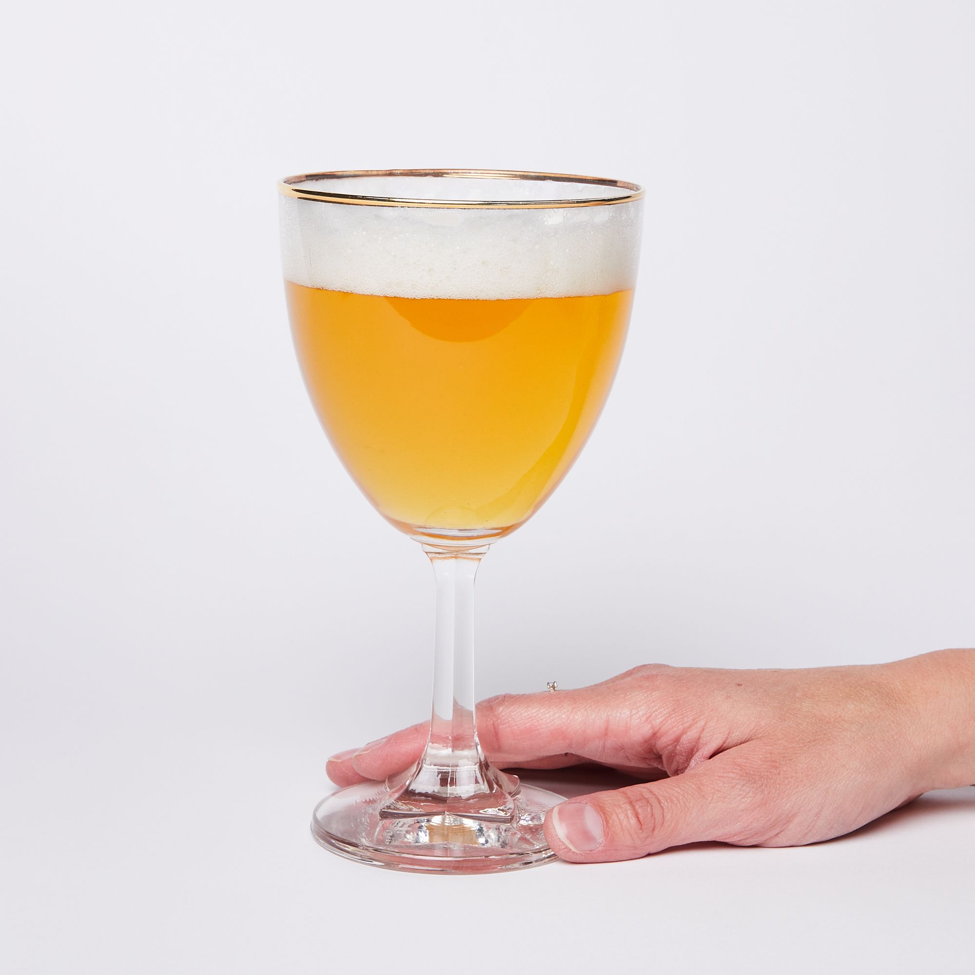 Hand holding the bottom of a stemmed clear beer glass with a gold rim full of beer with a thick layer of foam