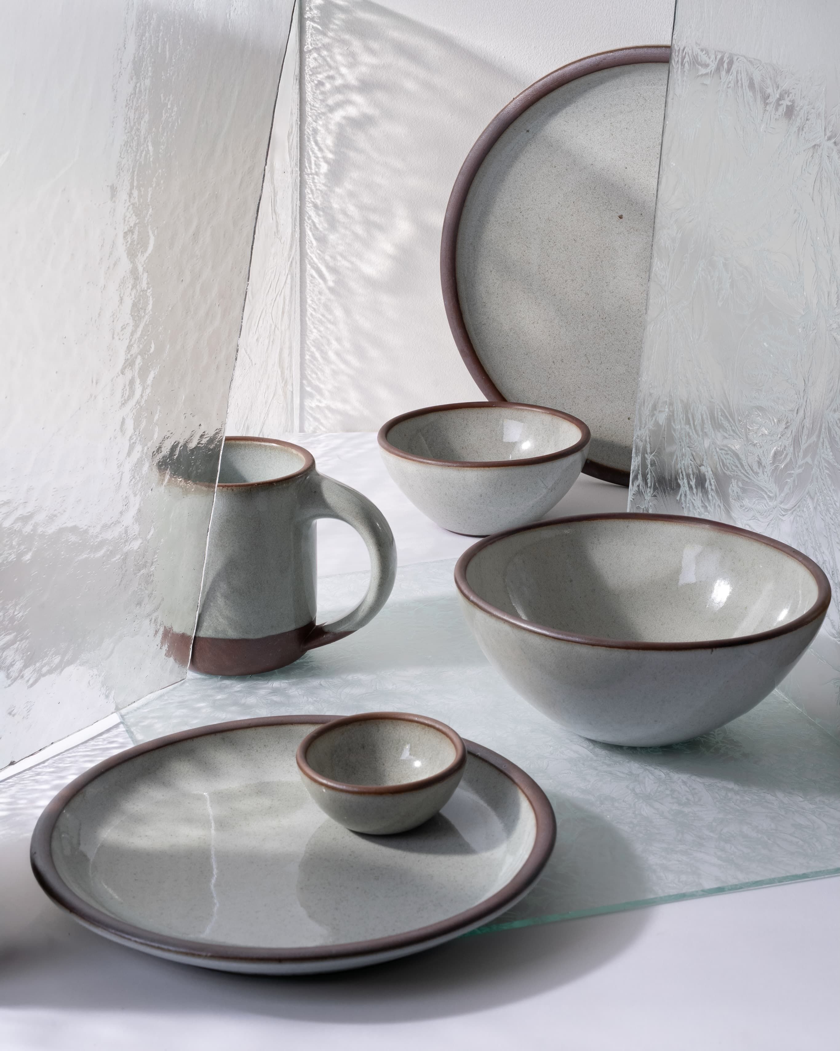 Collection of reflective icy white ceramic dinnerware with brown rims, including plates, bowls, and a mug, arranged with textured glass panels on a white surface.