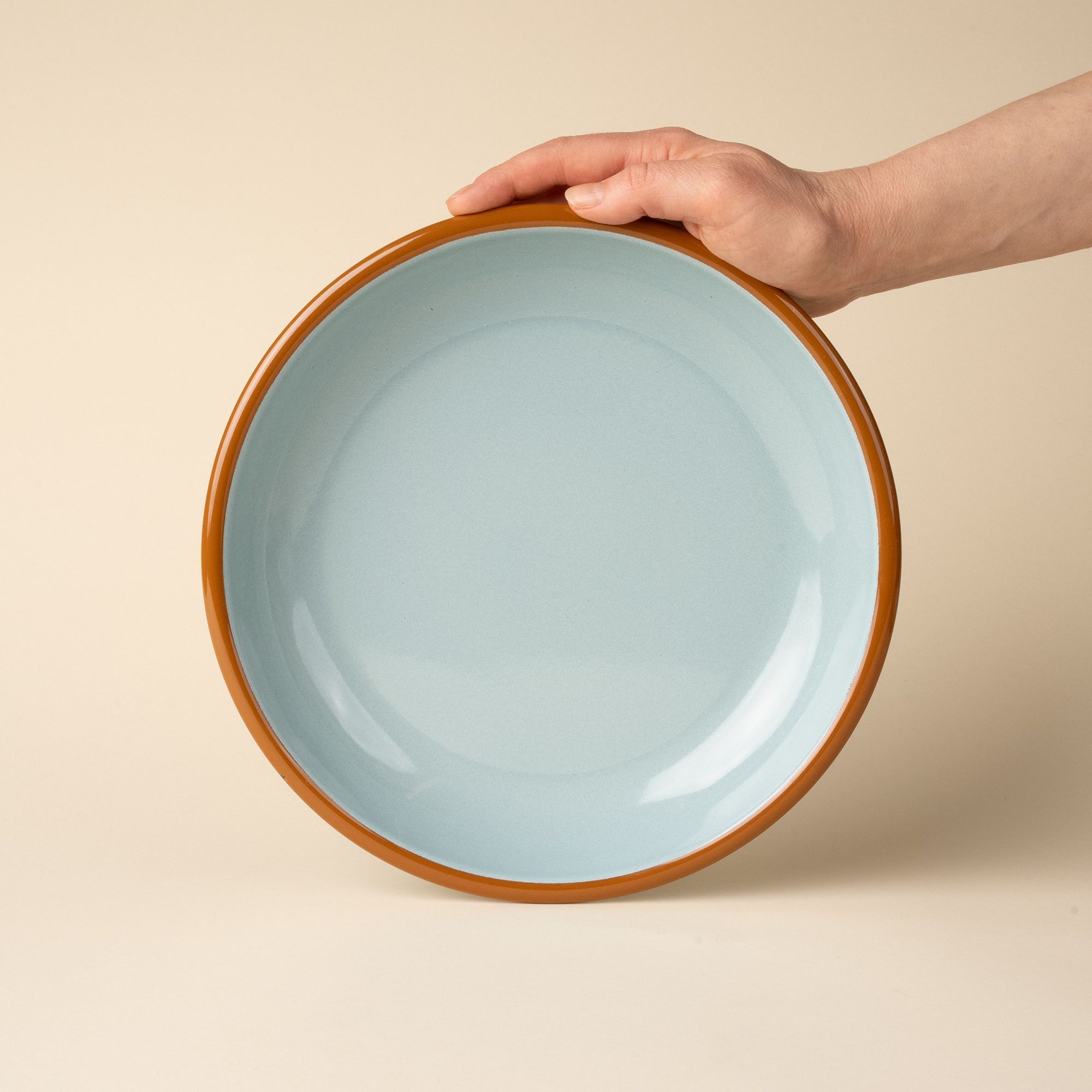 A hand props up an enamel dinner plate in robin's egg blue with a warm brown rim.