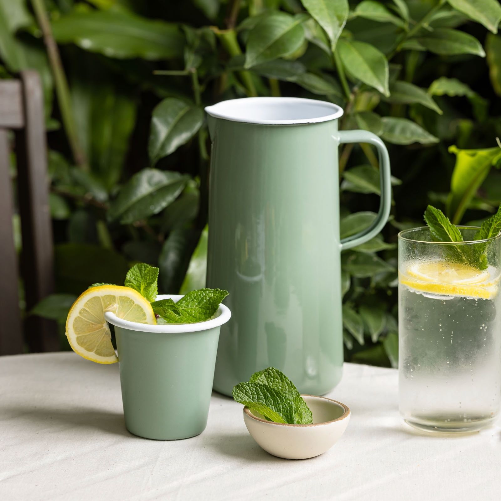 On an outdoor table, sits an enamel pitcher and cup in a soft sage green color. The cup is topped with mint and a lemon slice. Nearby is a glass of water with lemon and a tiny bowl filled with mint leaves.