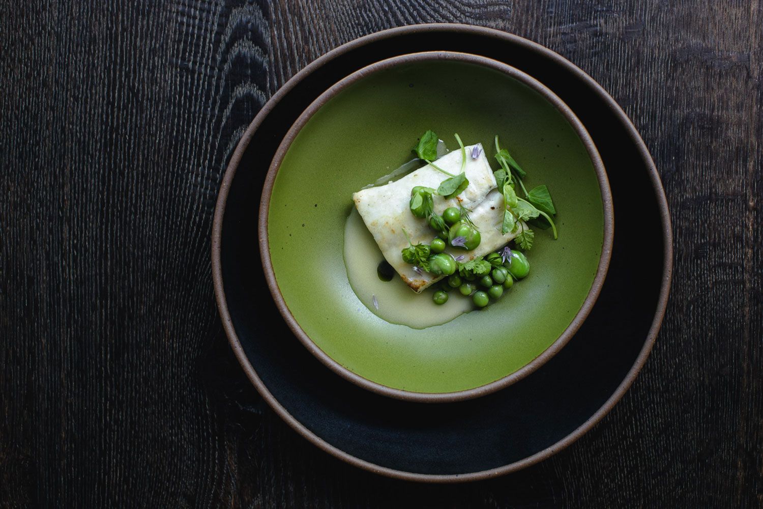 A sophisticated fish dish sits on a ceramic plate in a fern green color, on top of a black plate.