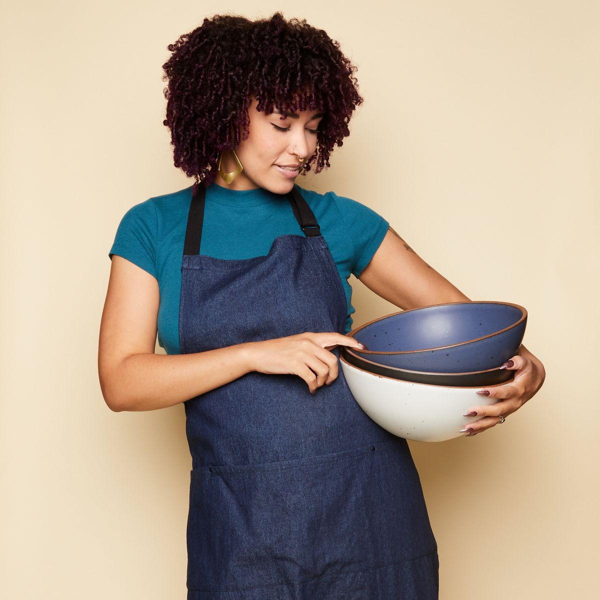 Woman looking down and smiling wearing a blue shirt with a denim blue apron over it and carrying 3 mixing bowls.