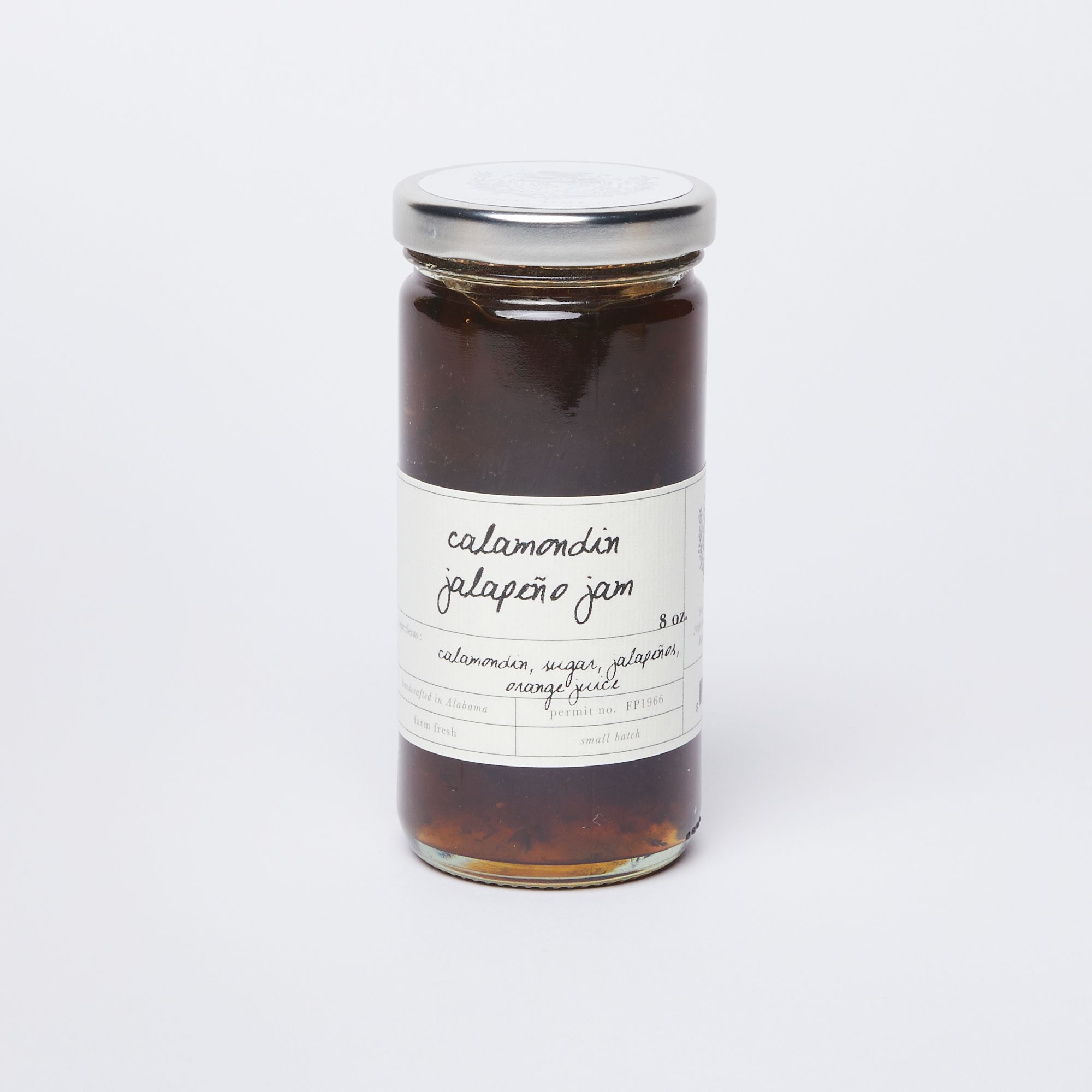 A clear glass jar full of amber colored jam with a silver lid and a white label that reads "Calamondin Jalapeño Jam"