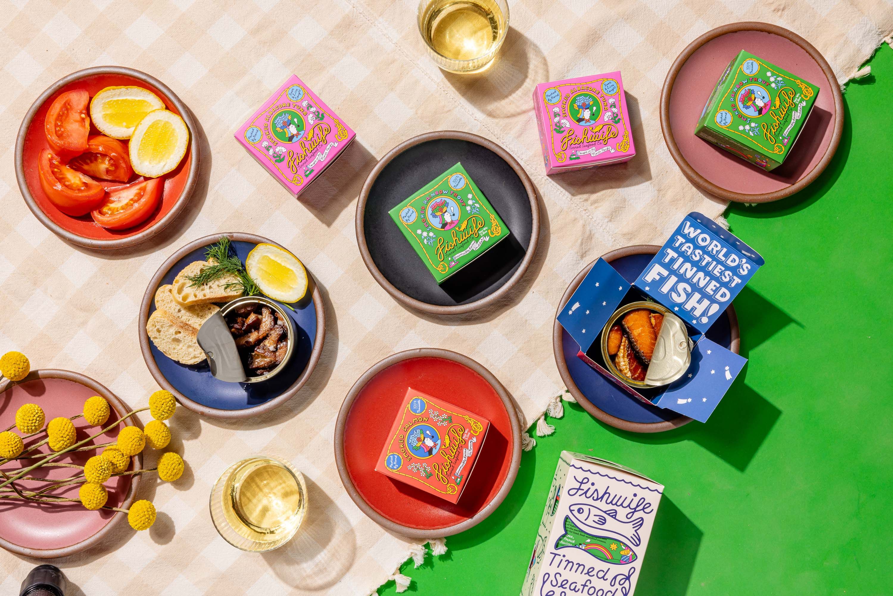 A spread of small ceramic plates in red, pink, blue and black colors, surrounded by illustrated tinned fish boxers in pink green and orange. On the plates are tinned fish, sliced bread, tomatoes, and lemons, and surrounded are flowers and wine in a glass.
