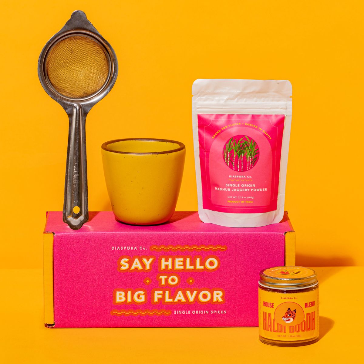 Pink box with a yellow Kulhad, strainer and tea on top. Image is set on a yellow background with a jar of haldi doodh in front of the pink box.