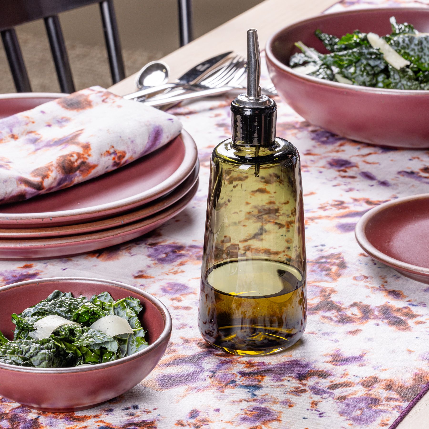 A short spruce green glass cruet on a table with a pink marbled tablecloth and plum ceramic dinnerware