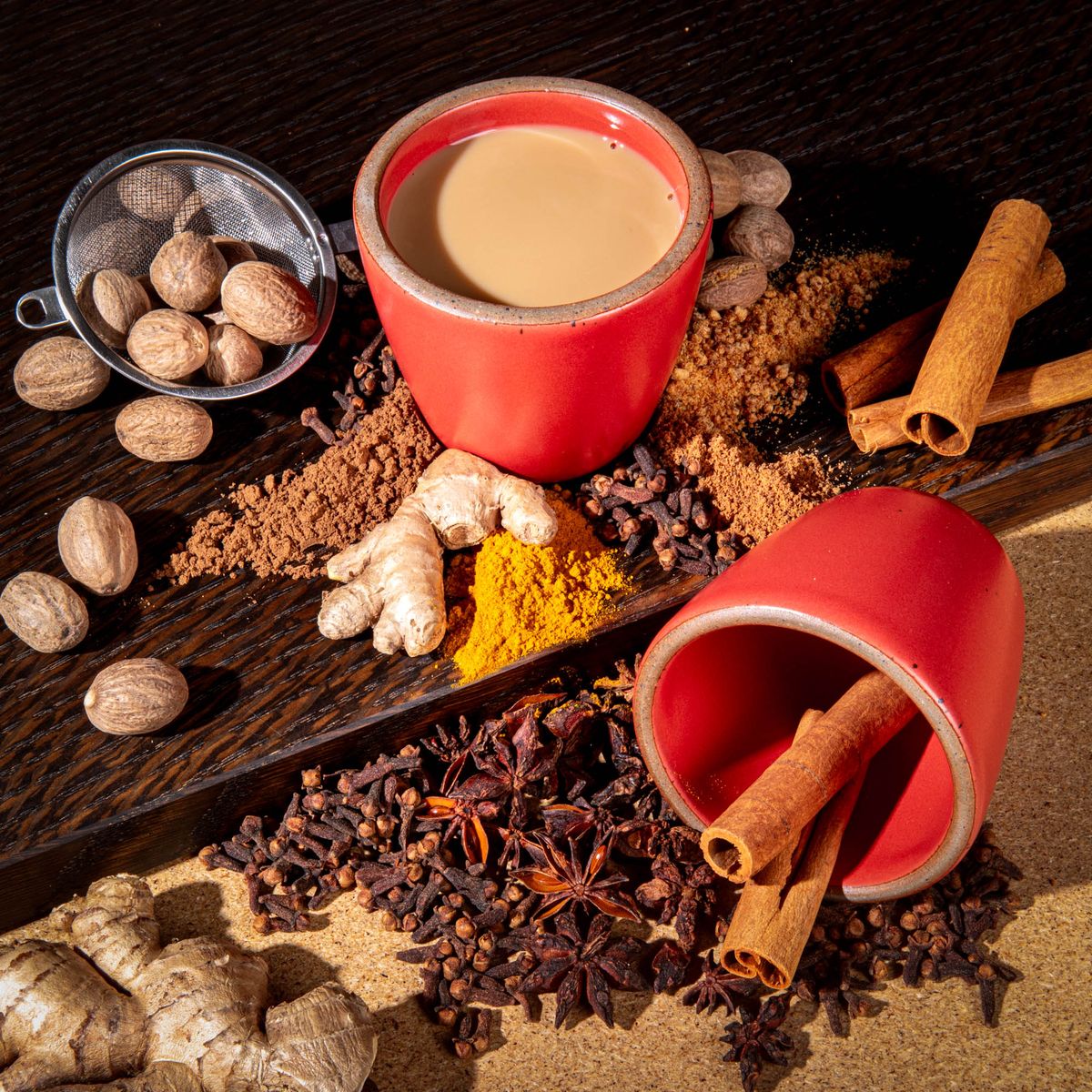 A short cup that tapers out to get wider at the top in a bold red color featuring iron speckles. The cup is filled with a buttered chai drink while another one is laying on its side holding cinnamon stick and surrounded by chai spices and a strainer. 
