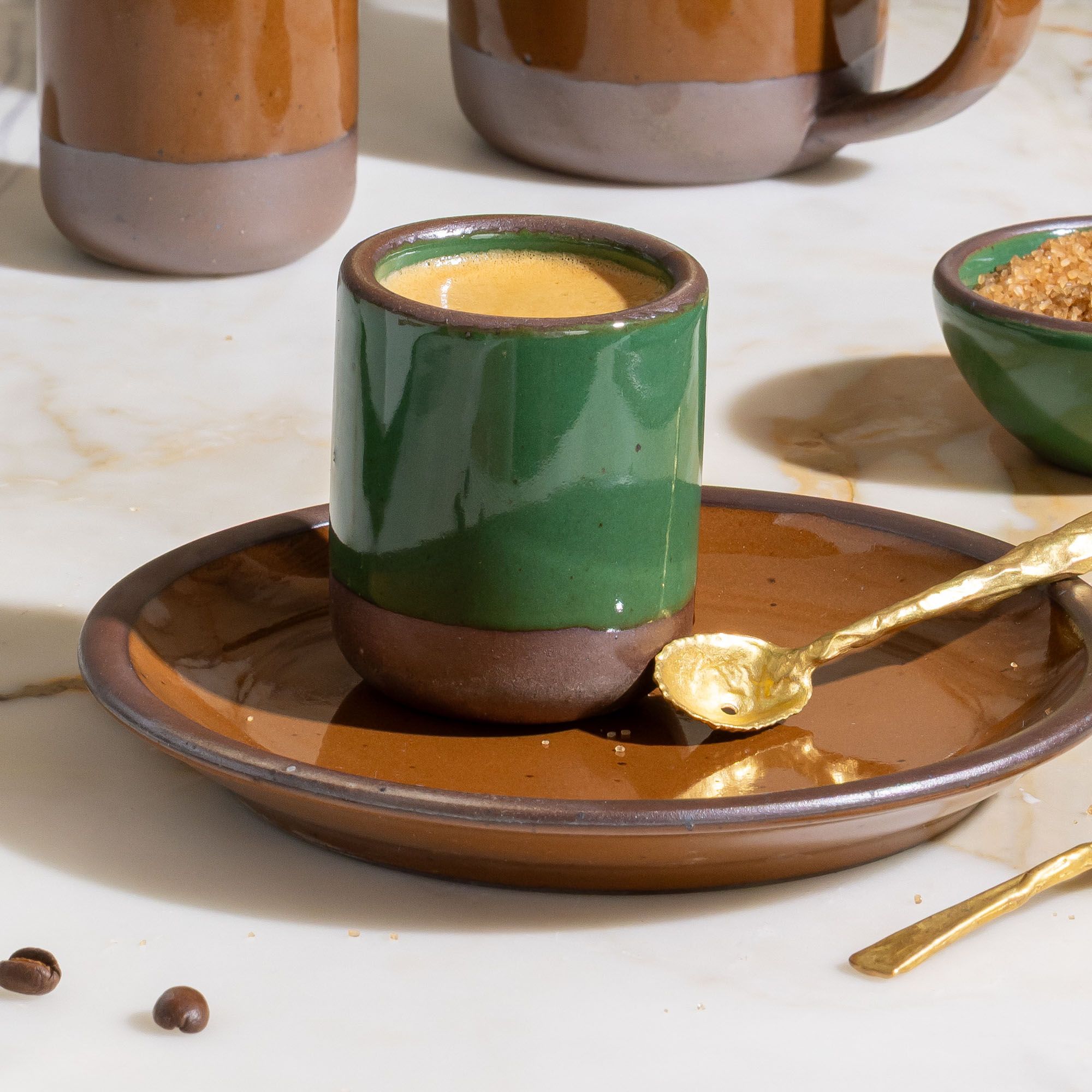 Glossy emerald green ceramic espresso cup with brown base on a brown plate, paired with a small gold spoon on marble surface.