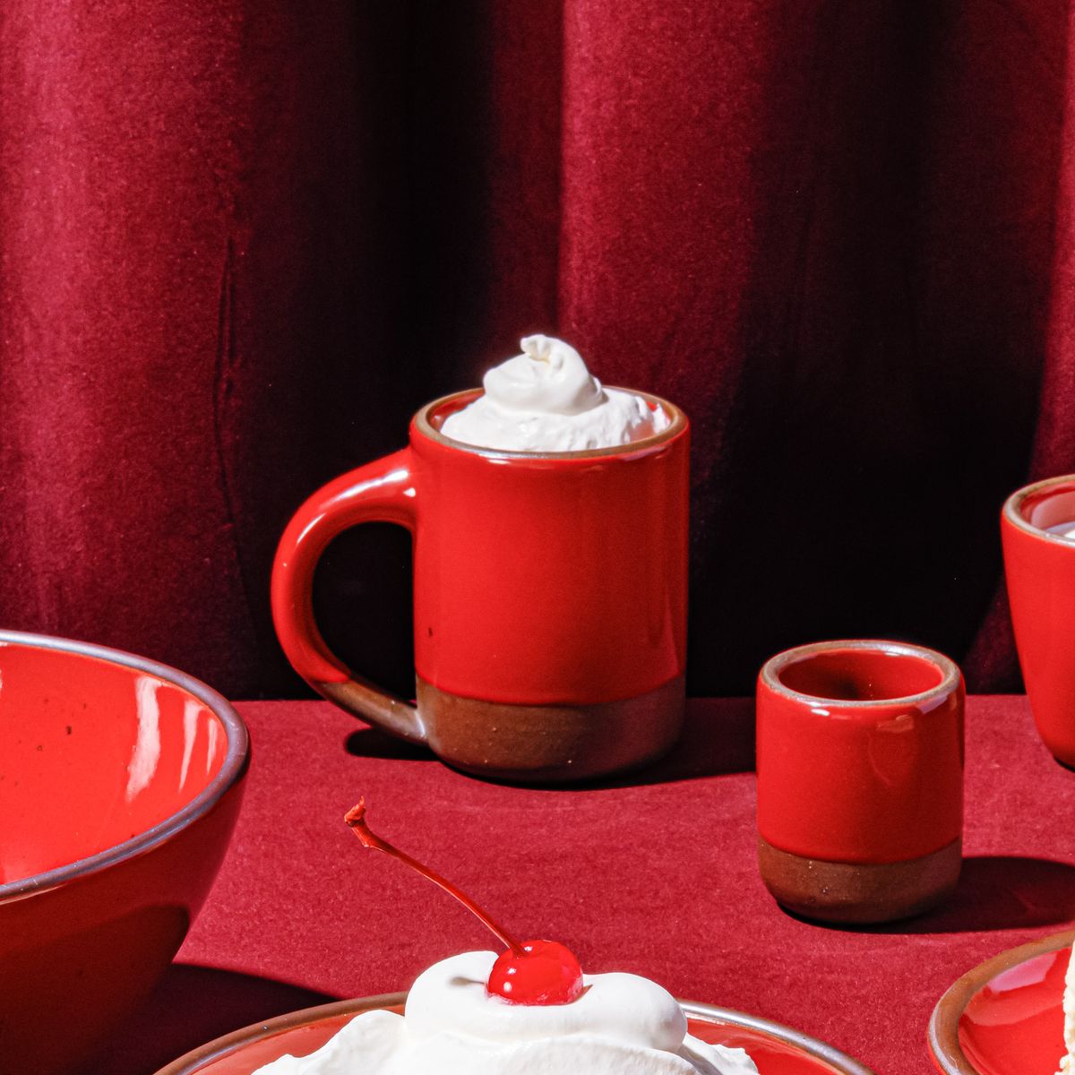 A glossy red ceramic mug topped with whipped cream, next to other ceramic forms and against a red fabric background.