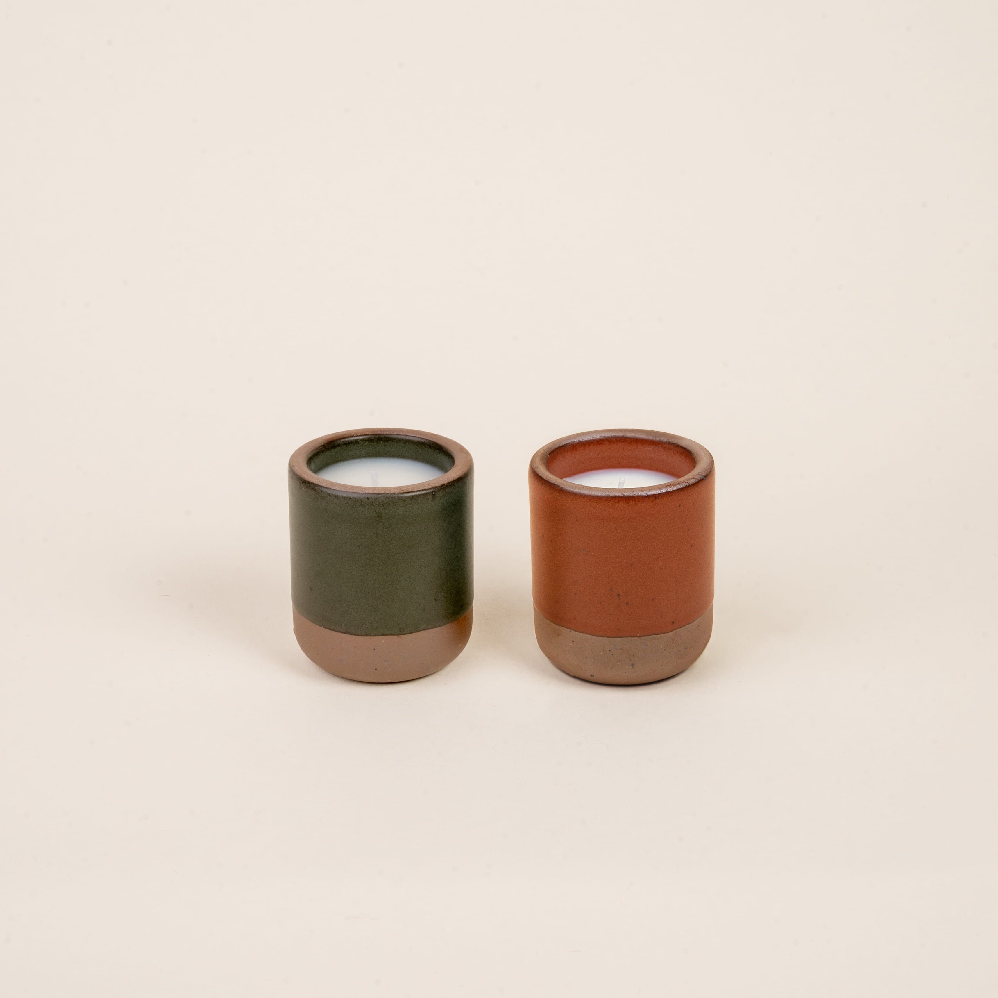 Two small ceramic vessels in forest green and cool terracotta colors with candles inside each.