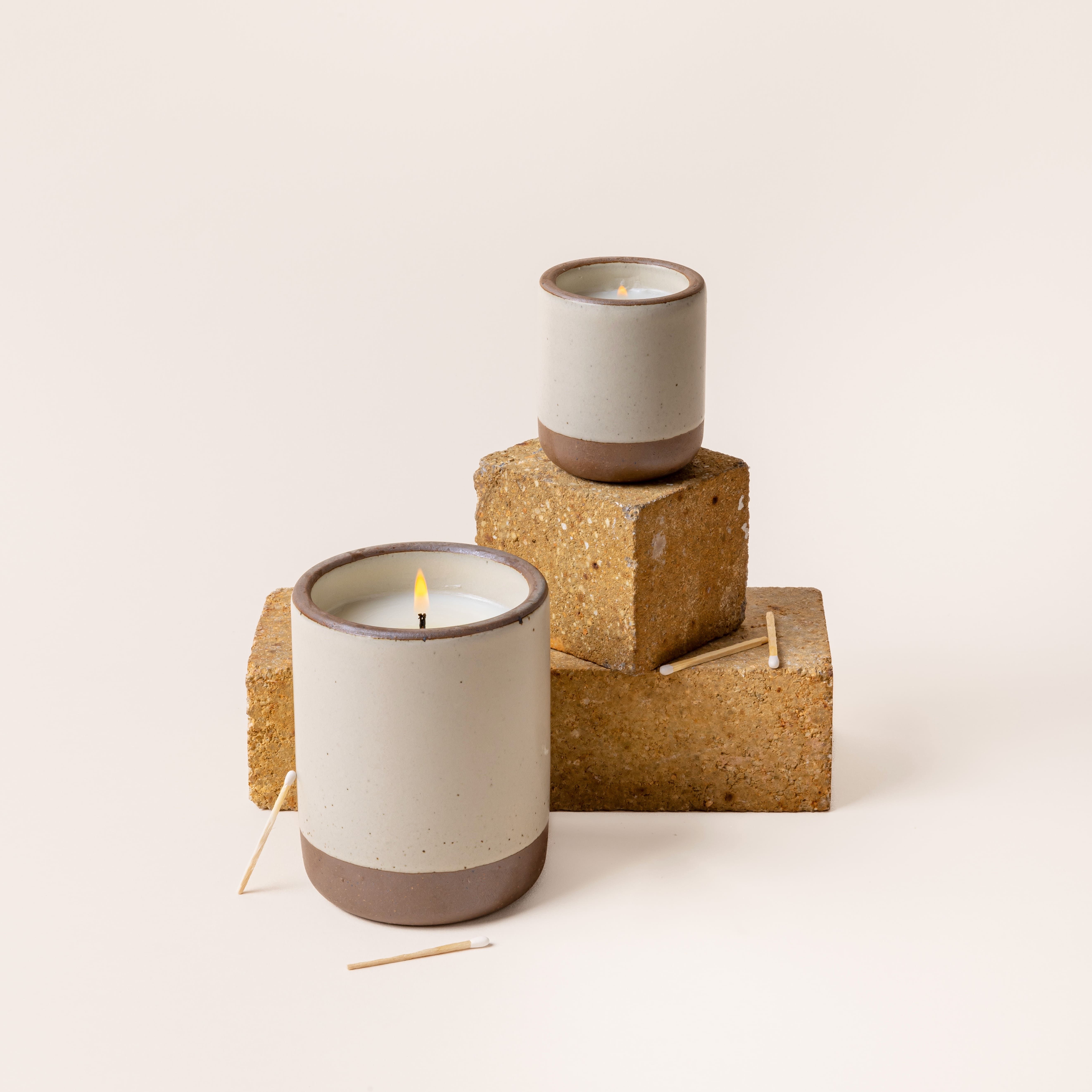 A large and small ceramic vessels in a warm, tan-toned, off-white color with candle inside each. The candles are lit and artfully arranged with brick and a few surrounding matches