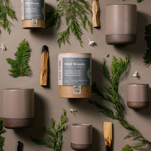 An artful layout of The Candle in Odd Woods - centered is a cardboard packaging tube with the candle's name, surrounded by small and large ceramic candles in a cylindrical vessel in a bold muted natural color, surrounded by sliced palo santo, cypress, and tiny flowers.