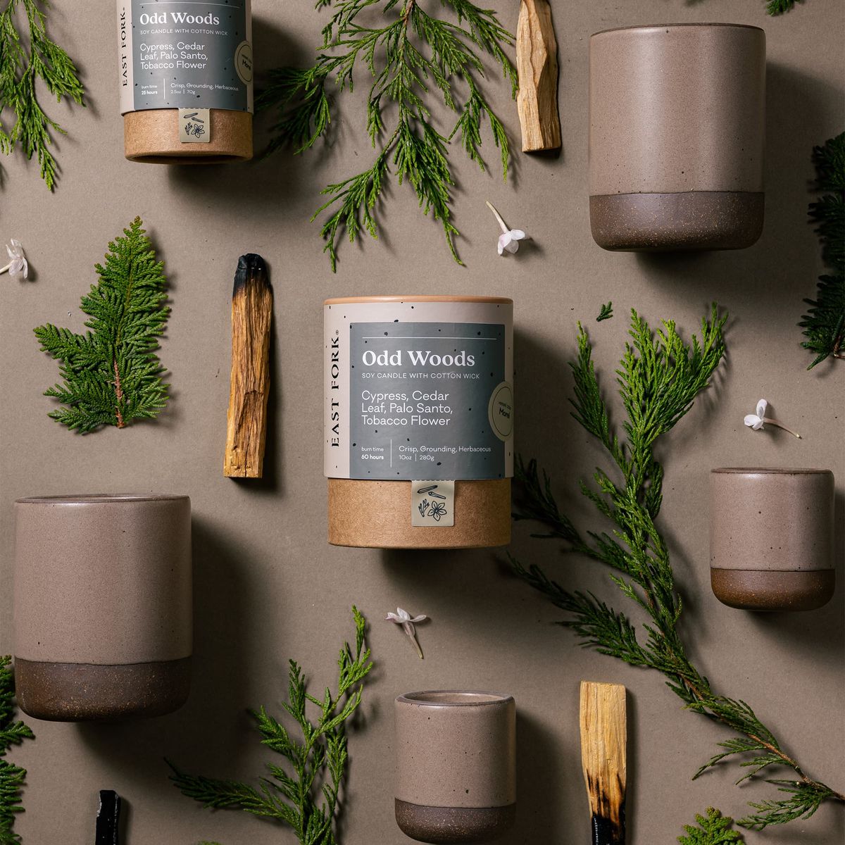 An artful layout of The Candle in Odd Woods - centered is a cardboard packaging tube with the candle's name, surrounded by small and large ceramic candles in a cylindrical vessel in a bold muted natural color, surrounded by sliced palo santo, cypress, and tiny flowers.