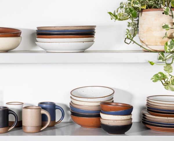 Two white shelves of ceramic plates and pots stacked in various colors: White, Off-white, Black, Terracotta, Beige, and Blue. Also on the shelf are ceramic mugs to the left, and a plant with trailing ivy on the top right.