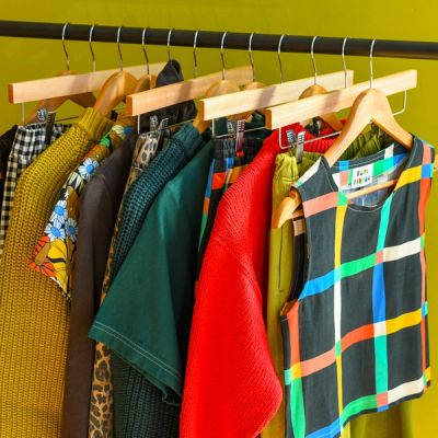 An image of a black rack with wooden hangers holding colorful clothing on a mustard green background