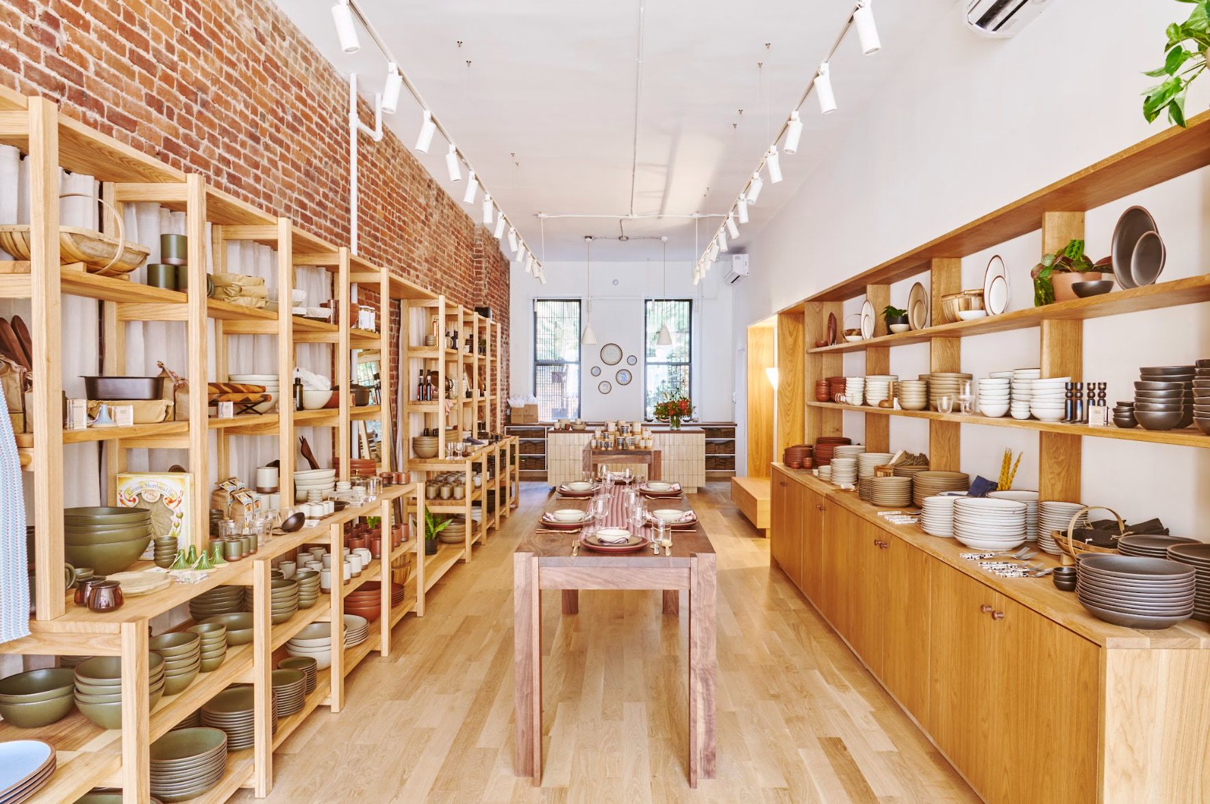 Inside the Brooklyn store location, with a brick wall on the left side and wood shelves lining the left and right walls. A table filled with place setting is in the middle leading to the cash wrap in the back.