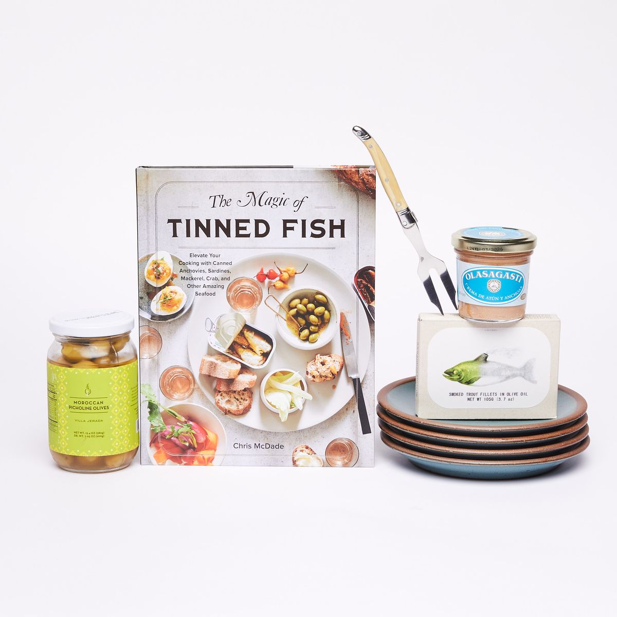 From left to right: Clear jar of olives with green and yellow label with white top, bookcover that reads "The Magic of Tinned Fish", an ornate two-pronged fork, four turquoise cake plates, a box with a picture of a fish, a glass jar with a white and blue label