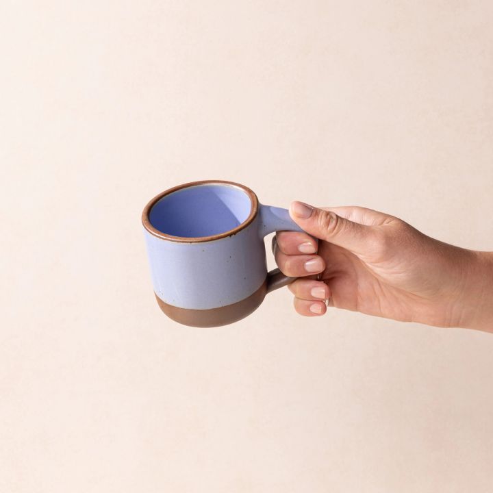 A hand holds a small sized ceramic mug with handle in a periwinkle featuring an unglazed rim