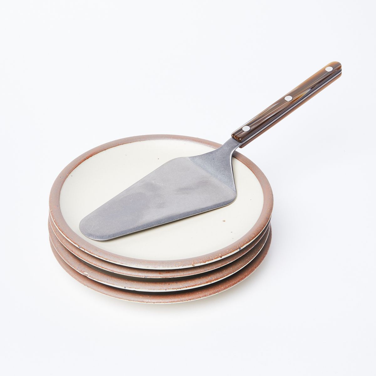 Silver Pie Server with a glossy brown handle with 3 silver circles on it sitting on top of a stack of 4 ceramic cake plates that are warm cream in color with a raw unglazed rim.