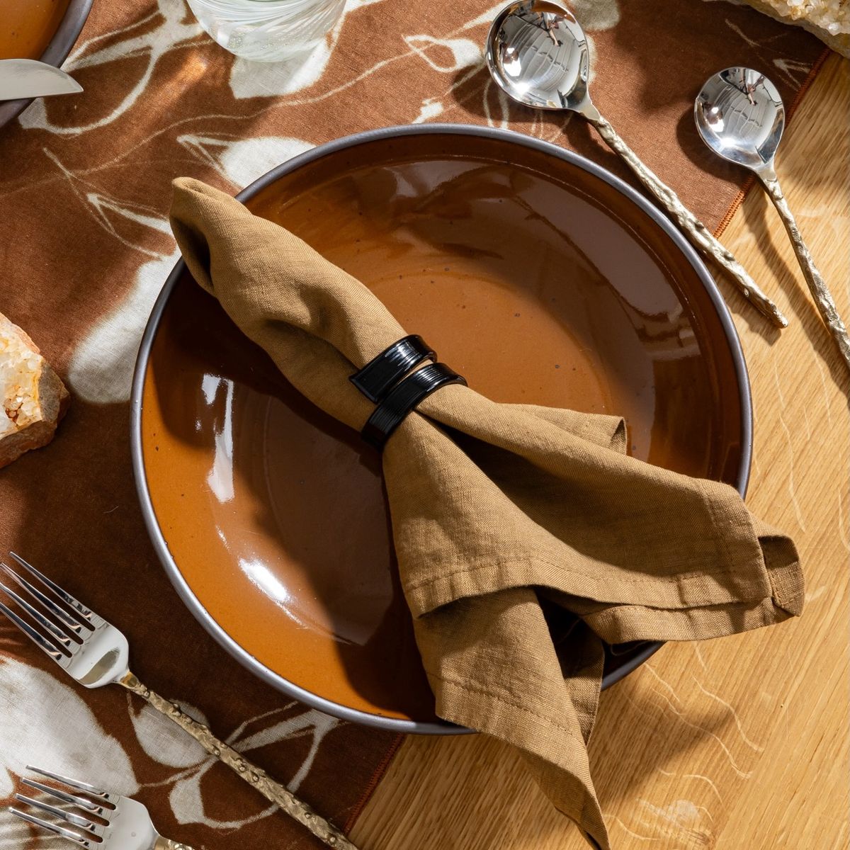 Brown linen napkin with amber glass napkin ring placed on a glossy brown glazed ceramic plate over patterned tablecloth.