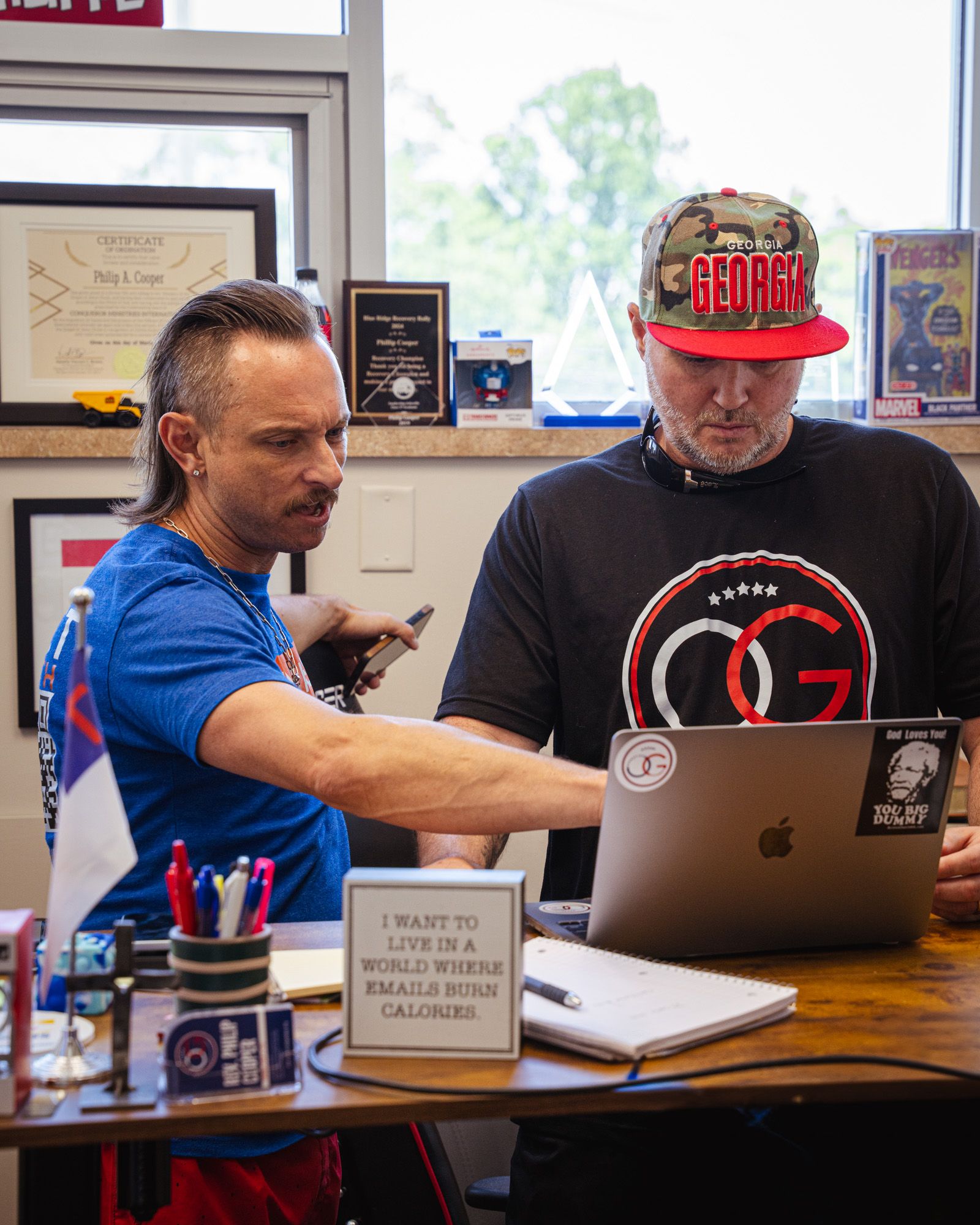 Two people are in an office standing, both looking and pointing at a laptop computer