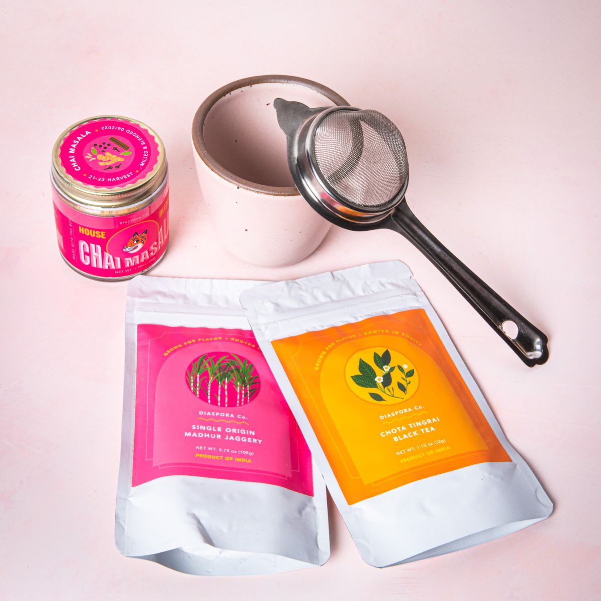 A chai bundle on a soft pink background featuring a soft light pink kulhad, a tea strainer, a pink jar of chai masala, and 2 packages of jaggery and black tea