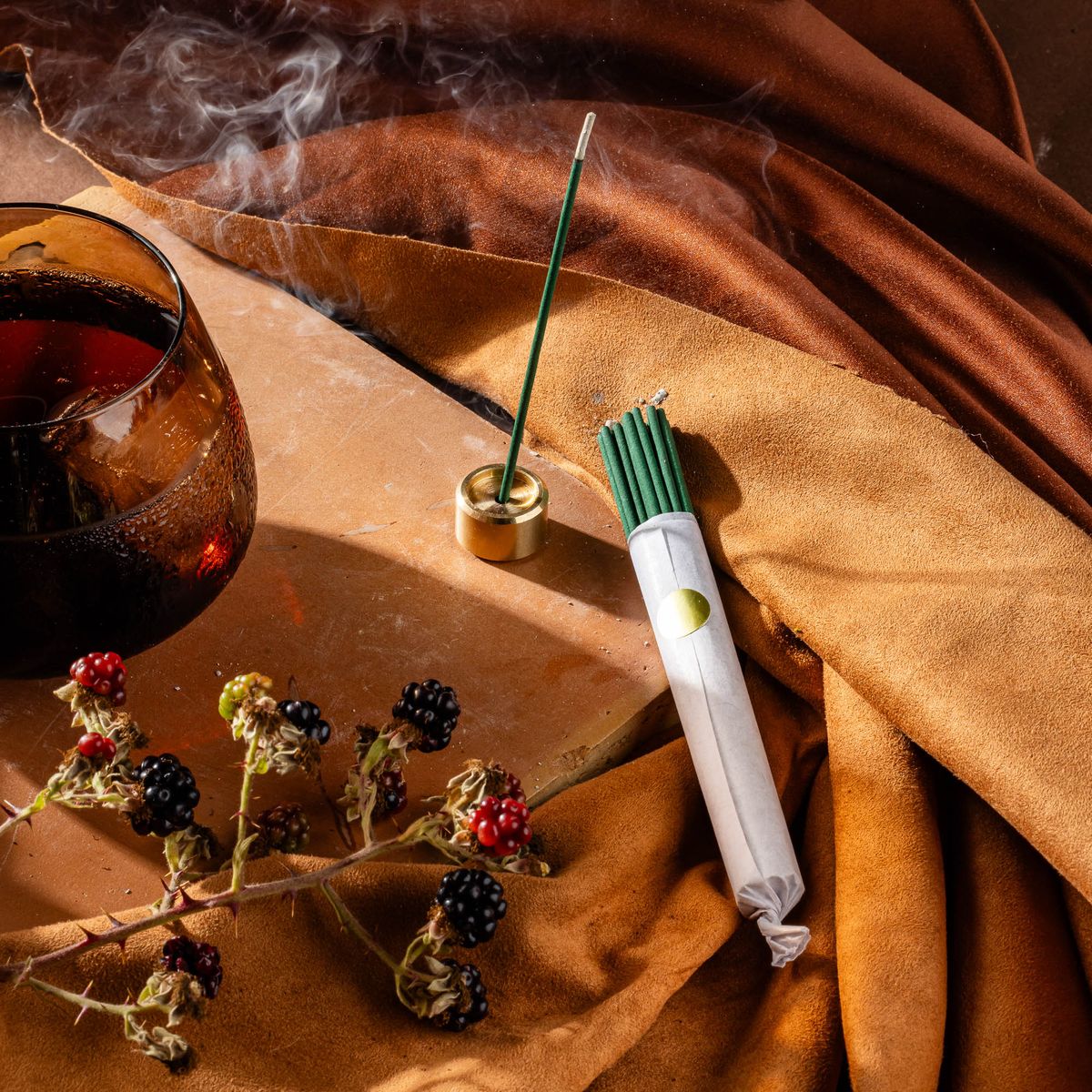 A bundle of teal incense sticks wrapped in paper sits next to a burning incense stick in a small brass holder. Both items are surrounded by a branch of berries and a whiskey snifter glass, on brown fabric.