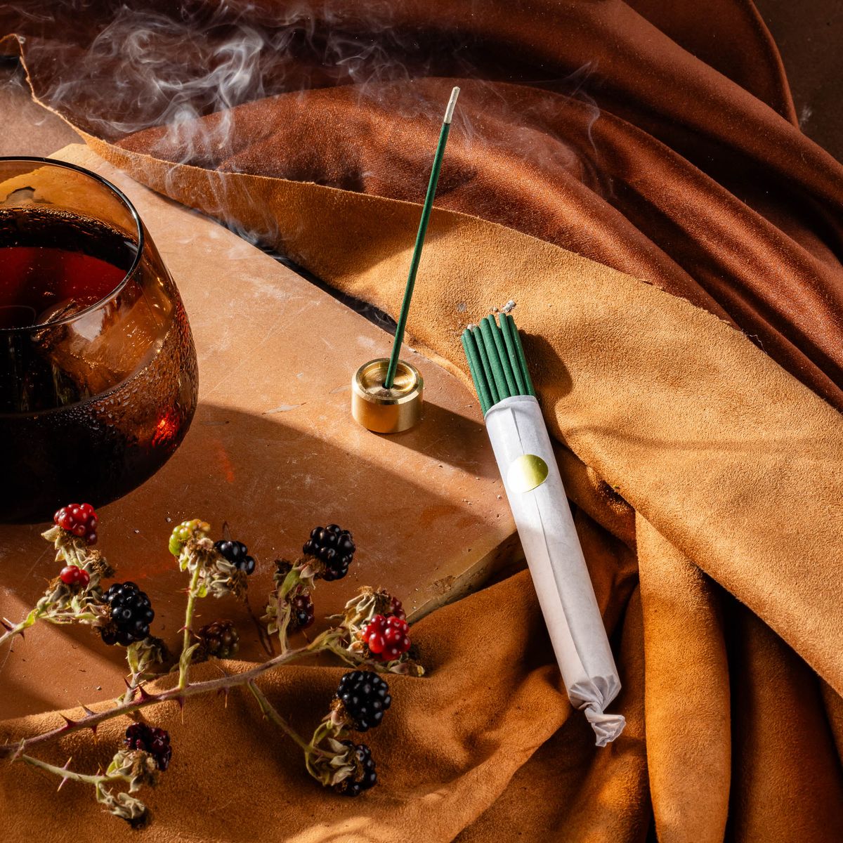 A bundle of teal incense sticks wrapped in paper sits next to a burning incense stick in a small brass holder. Both items are surrounded by a branch of berries and a whiskey snifter glass, on brown fabric. 