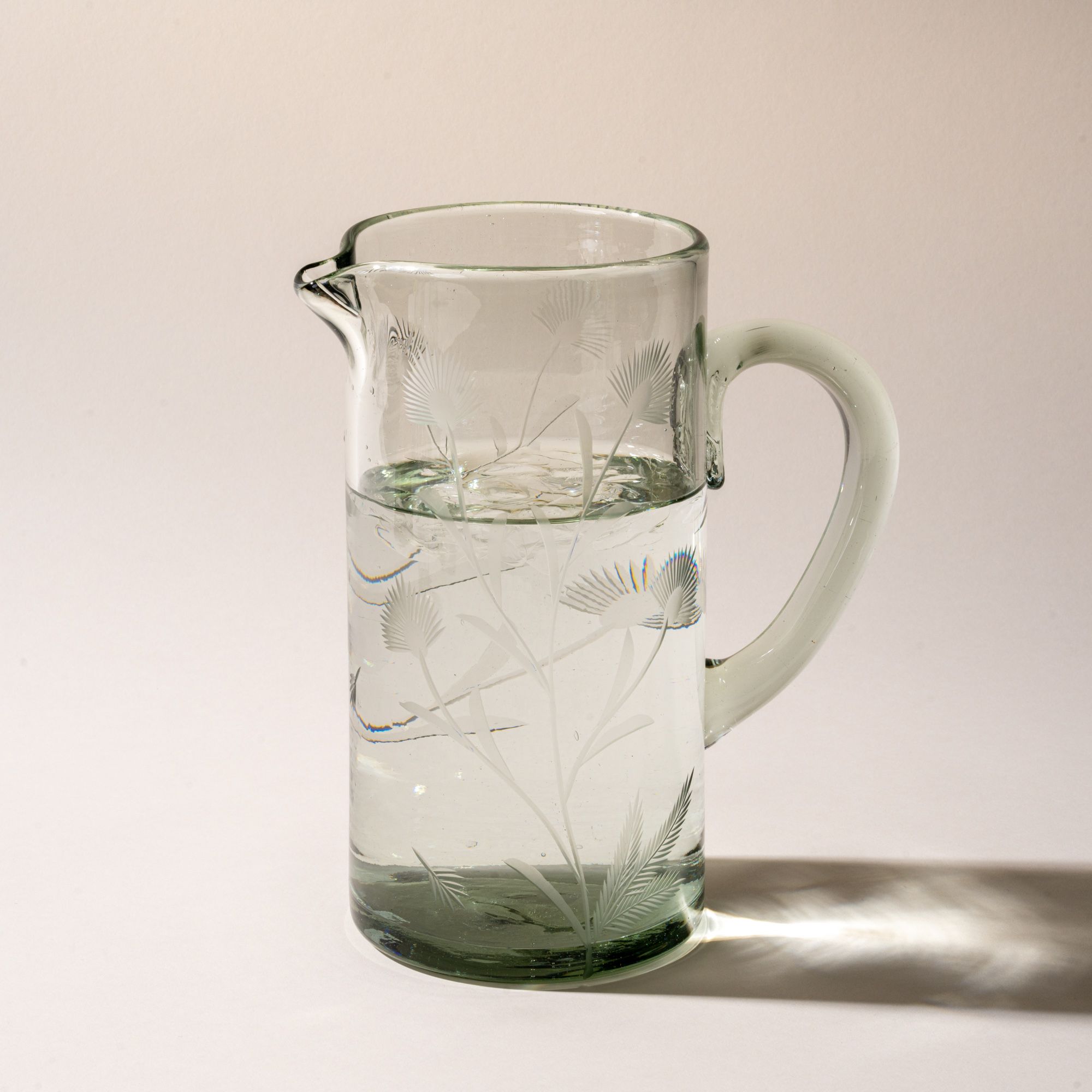 A large glass pitcher with handle made of recycled glass with etched thistle designs on it, and filled with water.