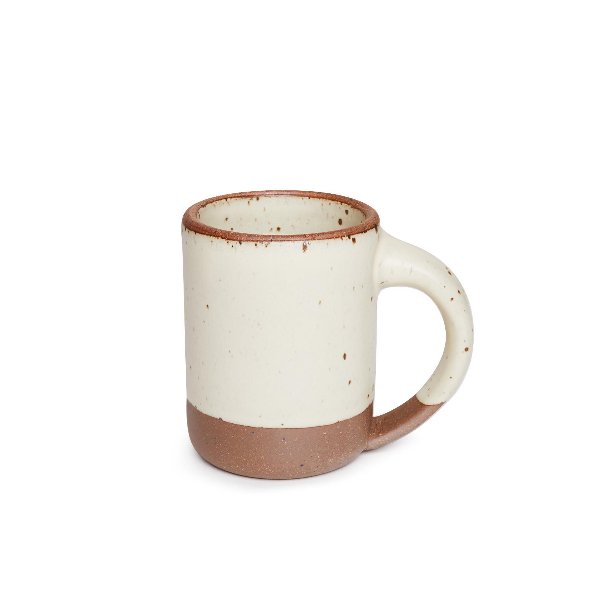 A medium sized ceramic mug with handle in a warm, tan-toned, off-white color featuring iron speckles and unglazed rim and bottom base.