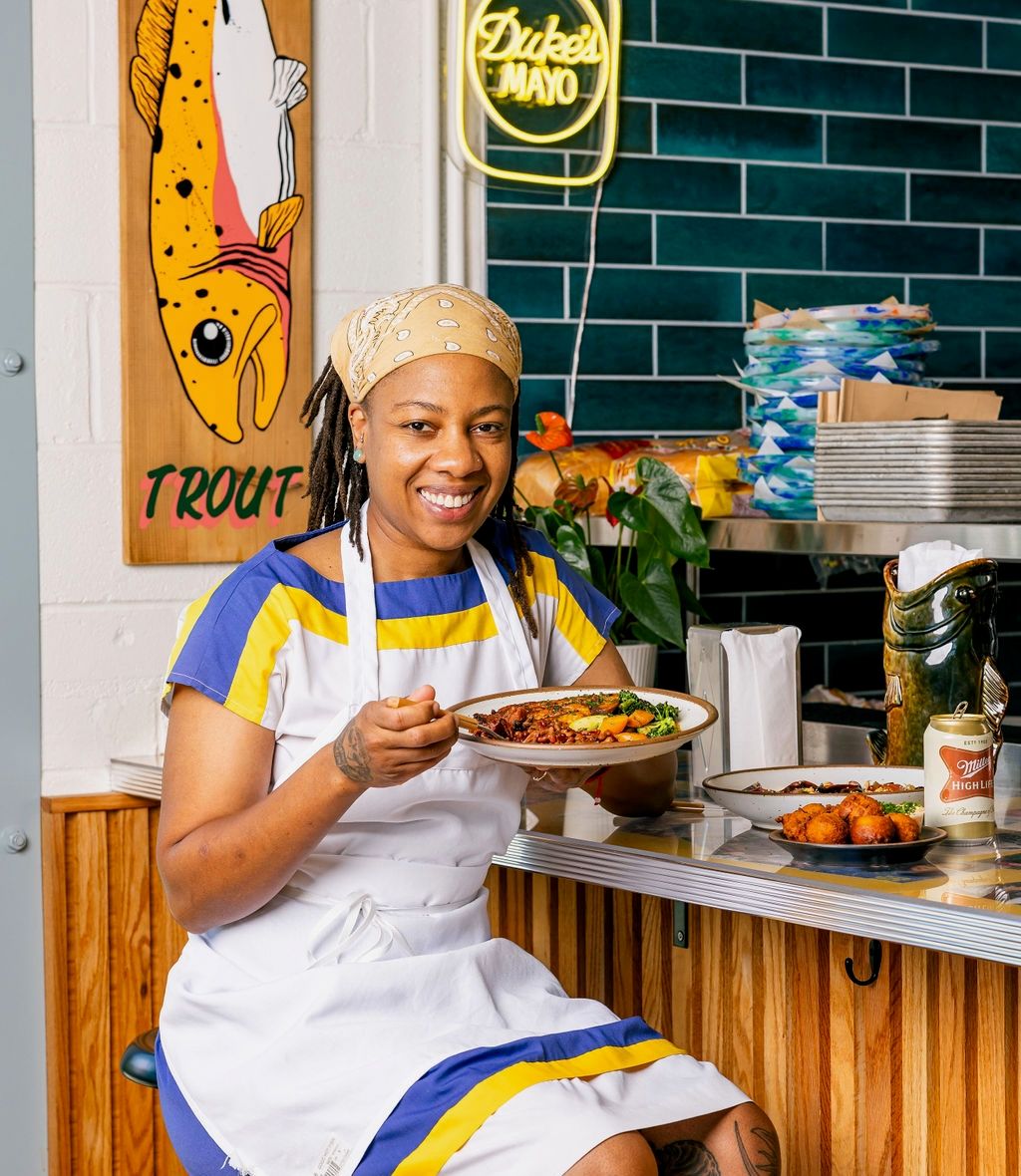 A woman in an apron sits at a restaurant counter holding a plate of food and smiling.
