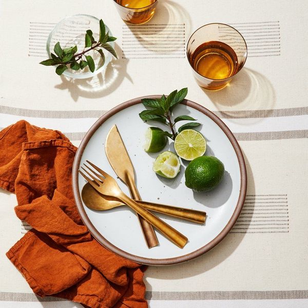 An overhead shot of a table setting. From the bottom, to the top: a natural colored tablecloth with a simple stripe pattern. A rumpled up burnt-orange-red napkin. An eggshell-colored stoneware dinner plate, adorned with brilliantly designed brass flatware, a sprig of mint, and limes. Nearby, a few glasses with water.